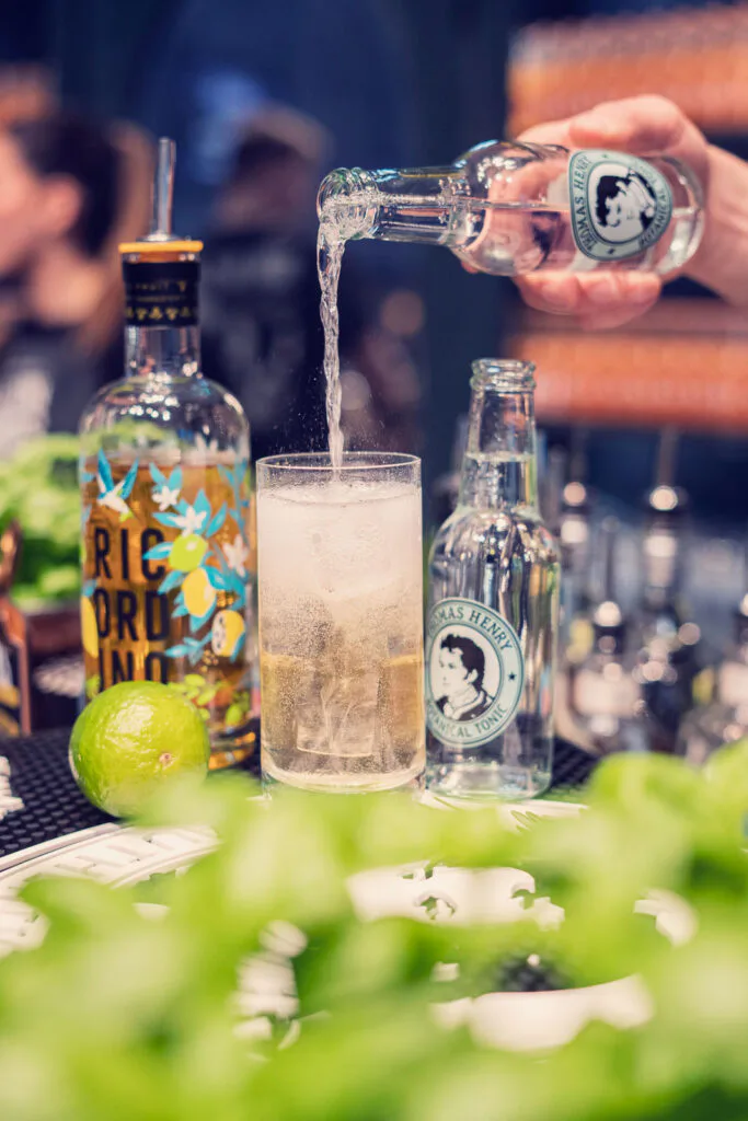 Eine Hand gießt aus einer klaren Glasflasche in ein hohes Glas an einer Bar; Limetten und Flaschen stehen daneben.