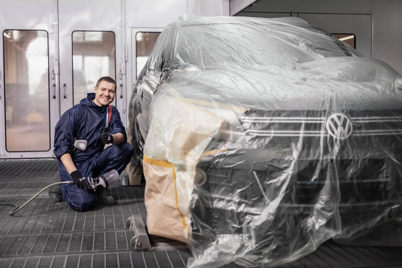 Mitarbeiter kniet mit Lackierpistole neben einem mit Folie abgedeckten VW in einer hellen Lackierkabine.
