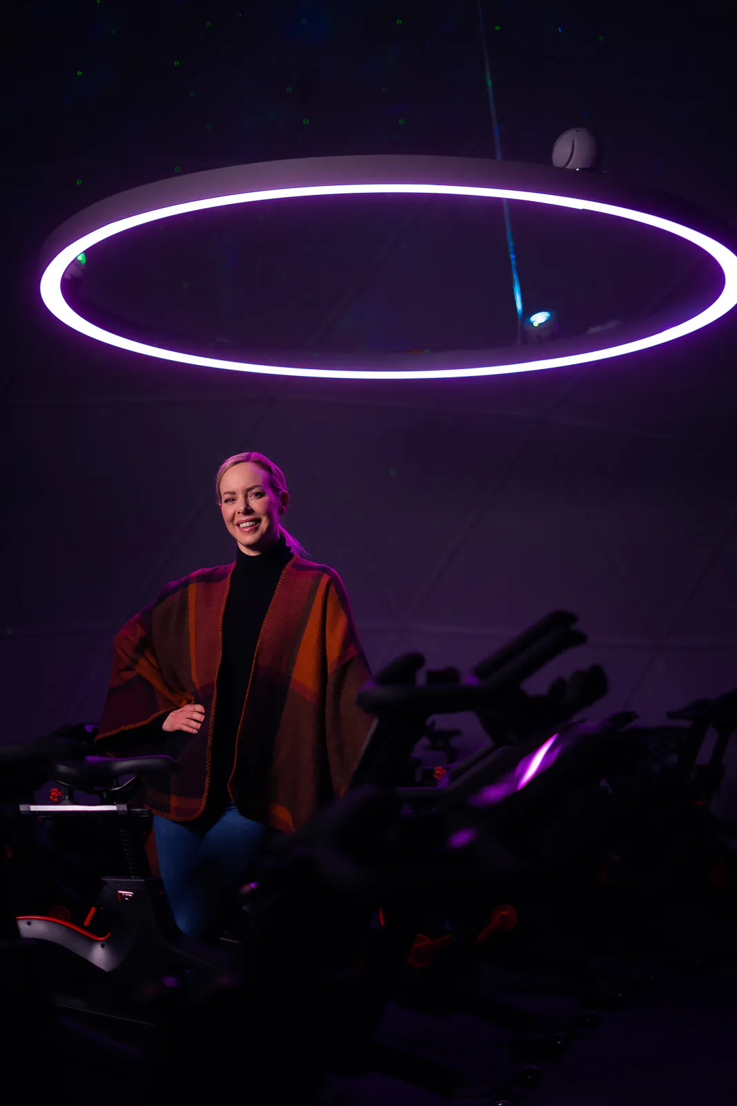 Isabel Edvardsson posiert zwischen Indoor-Bikes unter einem violett leuchtenden Deckenring.
