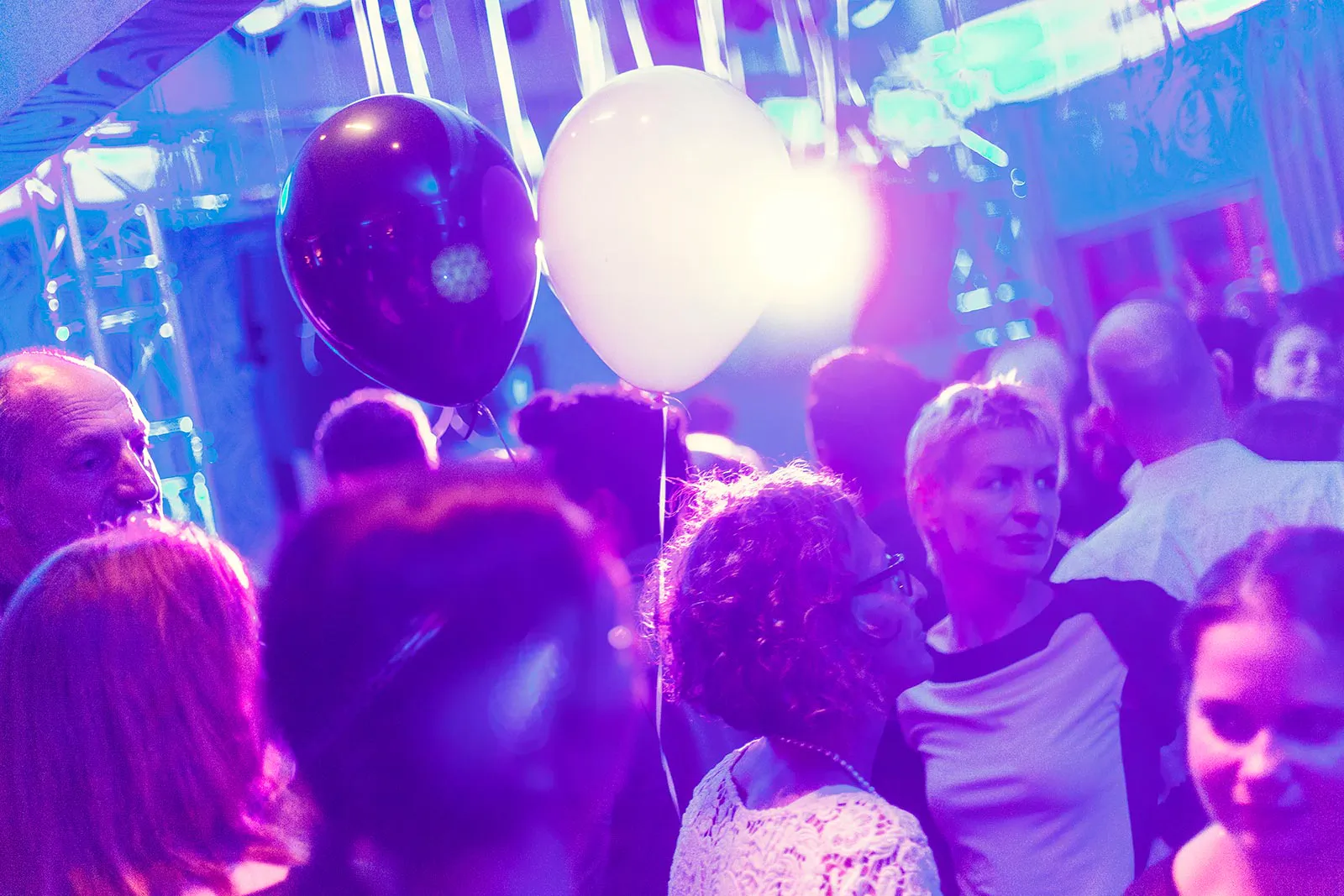Gäste stehen unter violett-blauem Licht mit schwarzen und weißen Luftballons im Veranstaltungsraum.