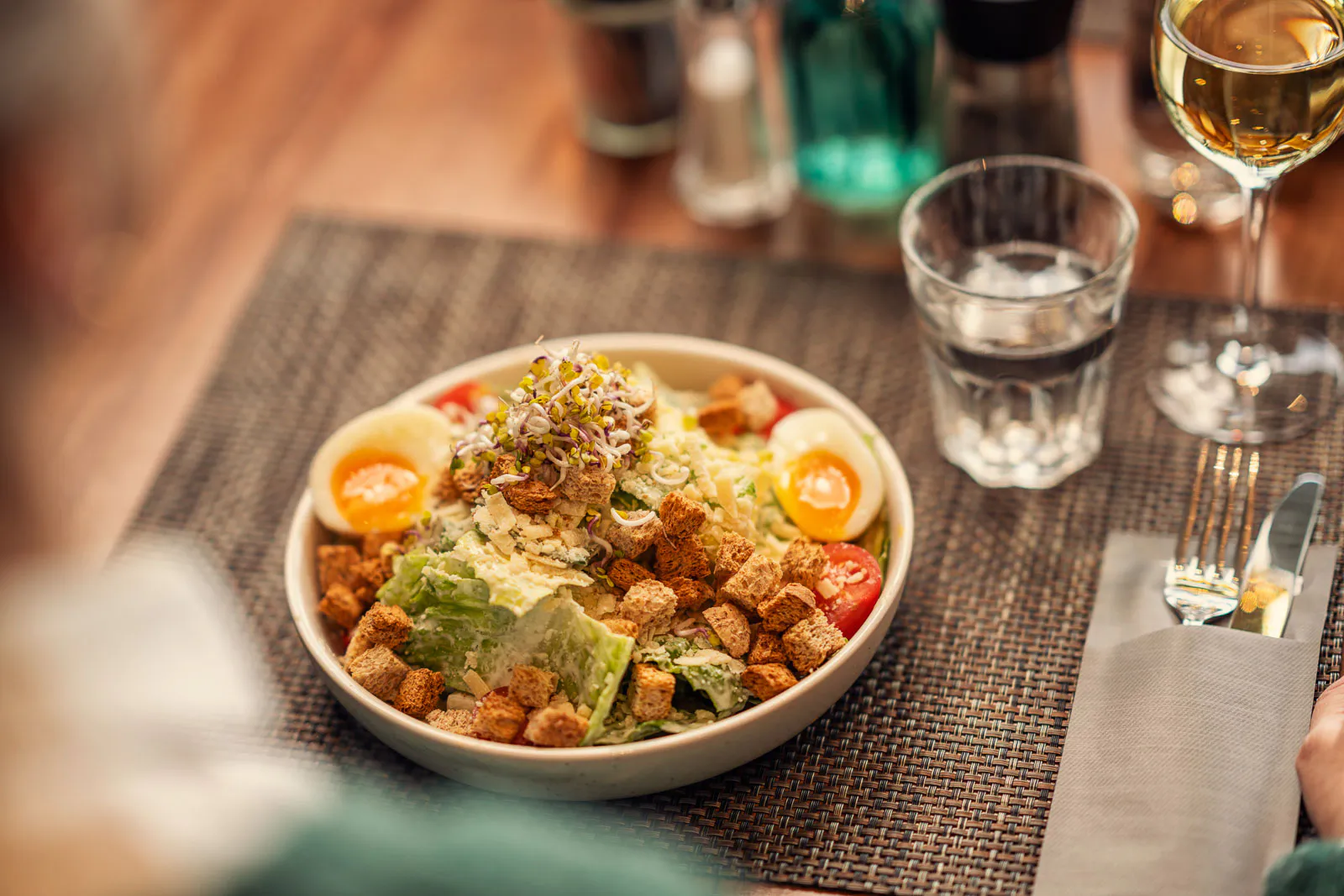 Salatschale mit Ei, Tomaten und weiteren Zutaten auf einem gedeckten Holztisch, daneben Gläser und Besteck.