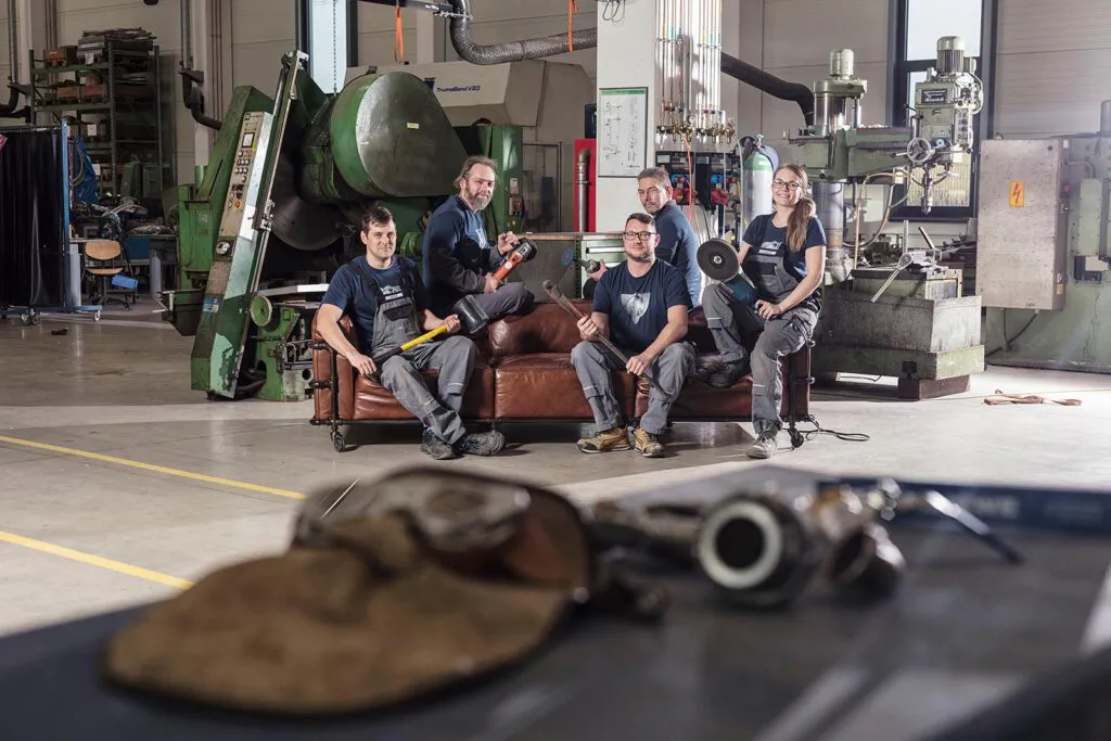 Fünf Personen mit Werkzeugen auf einem Ledersofa in einer hellen Werkhalle zwischen großen Industriemaschinen.