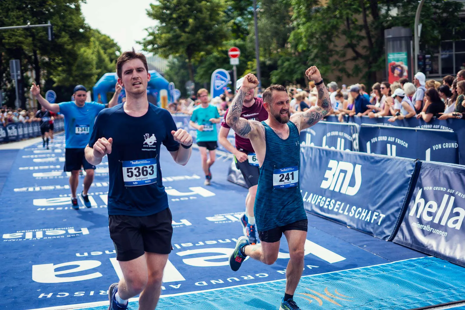 Läufer überqueren beim Hella Halbmarathon die Ziellinie, einer jubelt mit erhobenen Armen. Zuschauer säumen die Strecke.