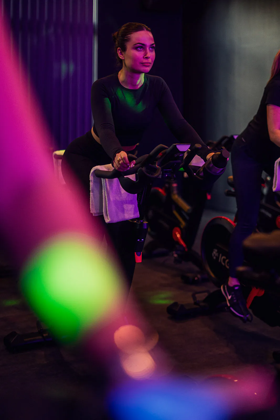 Frau in schwarzer Sportkleidung auf einem Spinning-Bike im Fitnessstudio bei violettem und grünem Licht.