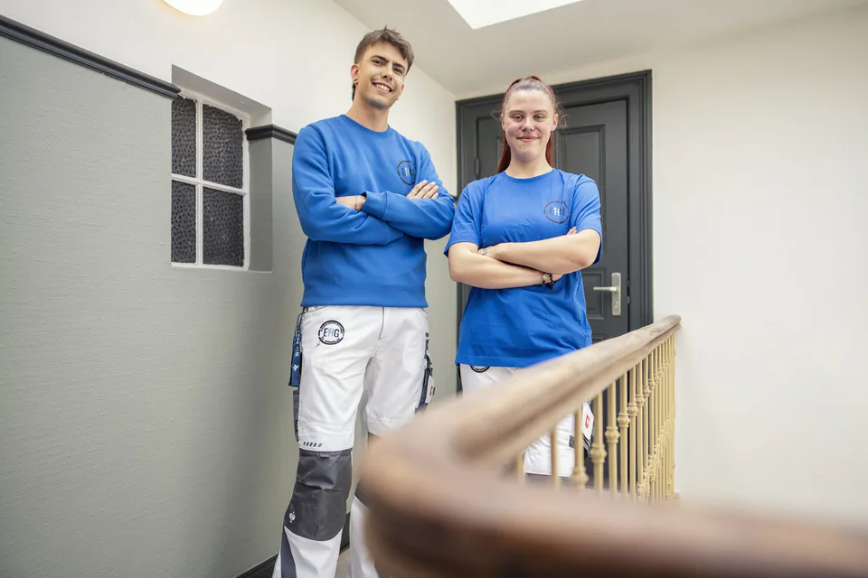 Zwei Handwerker in blauen Oberteilen und weißen Arbeitshosen stehen mit verschränkten Armen im hellen Treppenhaus.