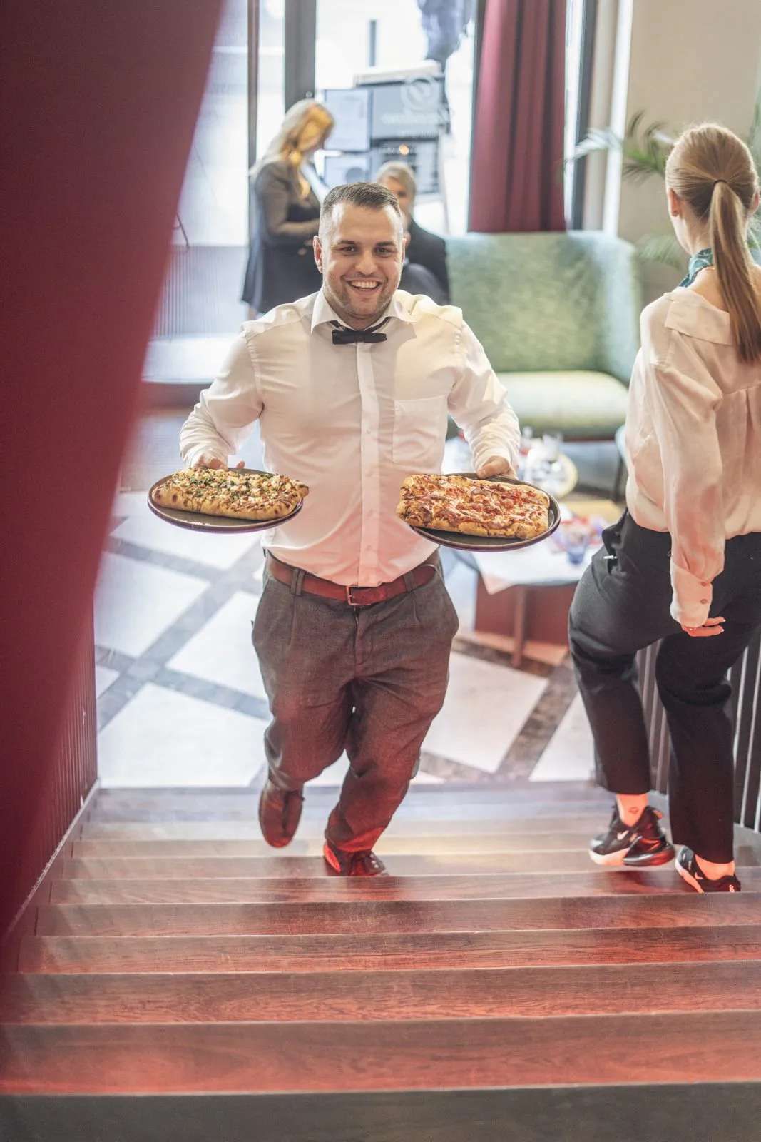 Lächelnder Mann mit weißem Hemd und Fliege trägt zwei Pizzen eine Treppe im Restaurant hinauf, neben ihm eine Frau.