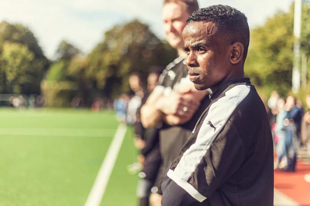 Zwei Männer in schwarz-weißer Sportkleidung stehen am Rand eines sonnigen Fußballplatzes.
