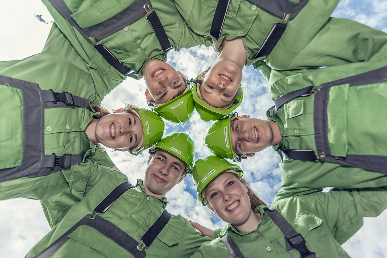 Personen in grüner Arbeitskleidung und Schutzhelmen blicken im Kreis lächelnd von oben in die Kamera.