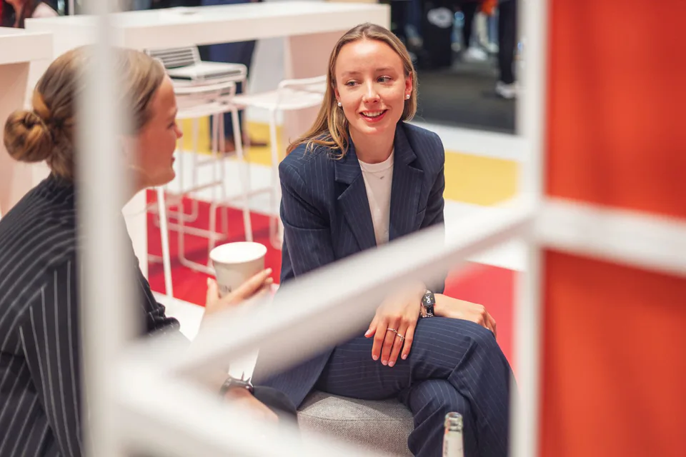 Zwei Frauen in dunklen Nadelstreifenanzügen sitzen im Gespräch am hellen Messestand, eine hält einen Pappbecher.