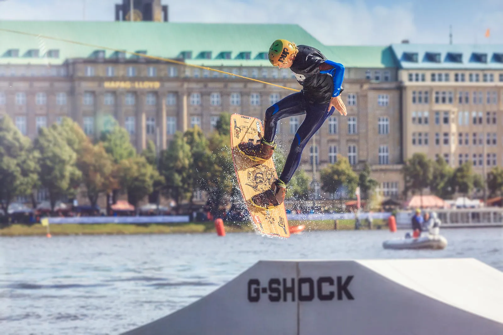 Wakeboarder mit Helm springt bei Sonnenlicht über eine G-Shock-Rampe auf einem Gewässer vor städtischer Kulisse.