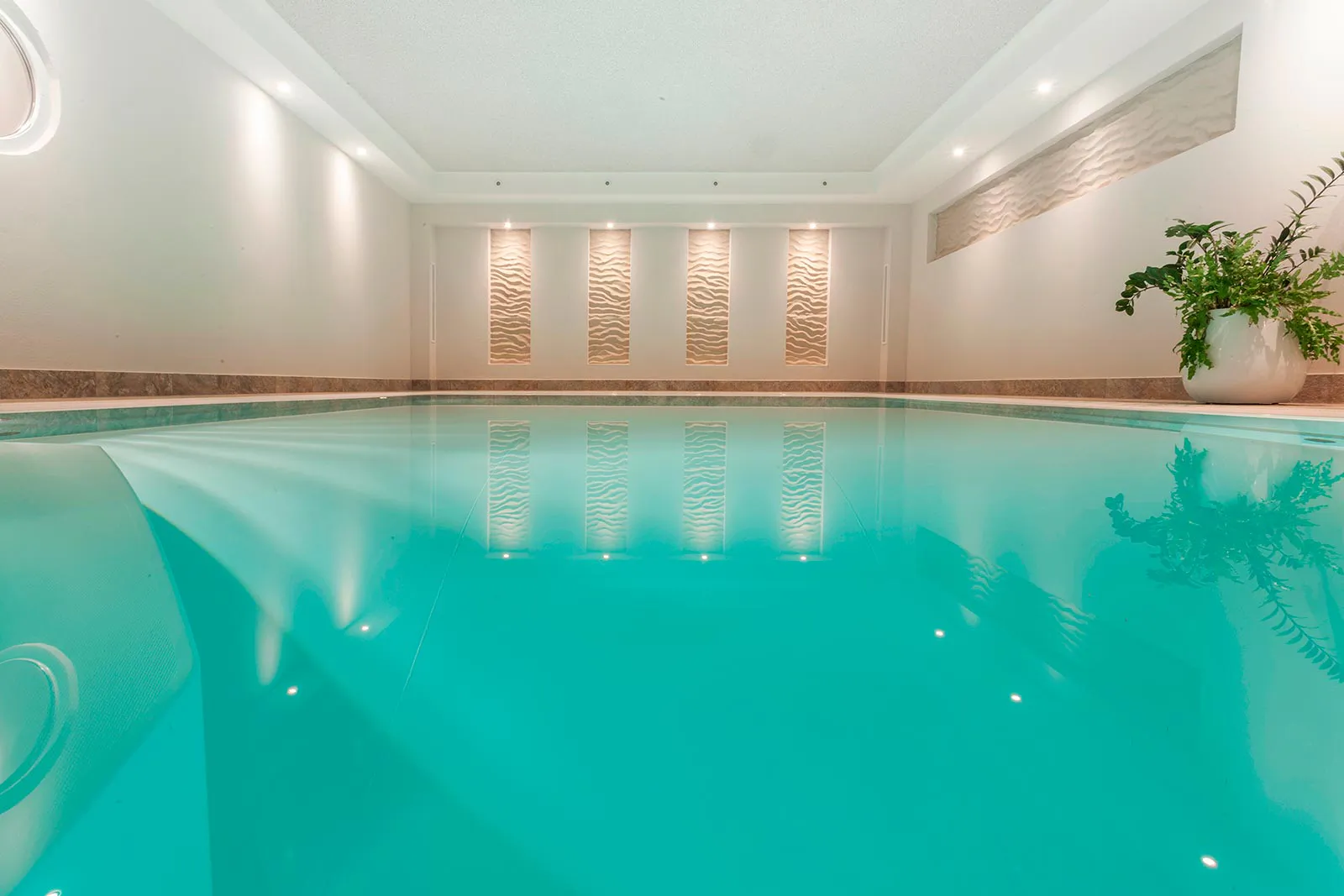 Hotel-Innenpool mit türkisfarbenem Wasser, Einstiegstreppe, beleuchteten Wandreliefs und einer Pflanze am Beckenrand.