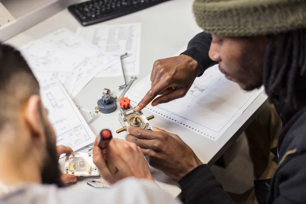 Zwei Personen arbeiten mit Schraubendreher an einem technischen Bauteil auf einem Tisch mit Zeichnungen.
