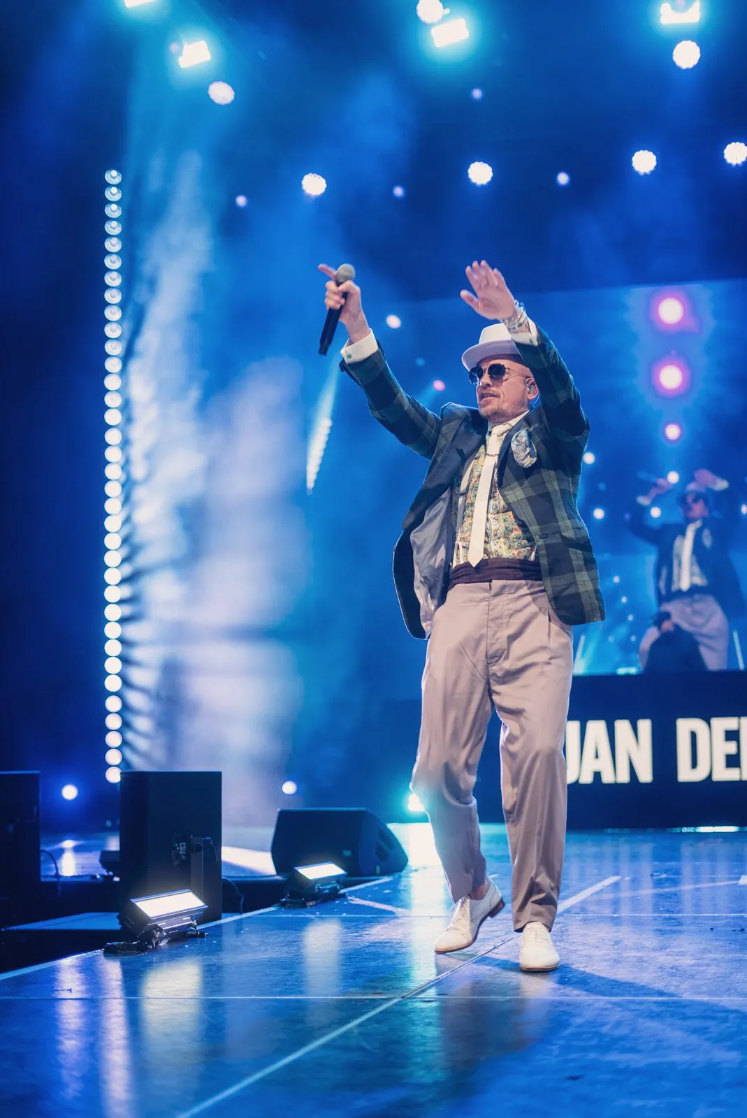 Jan Delay mit Hut, Sonnenbrille und Mikrofon, die Arme erhoben auf einer blau beleuchteten Konzertbühne.