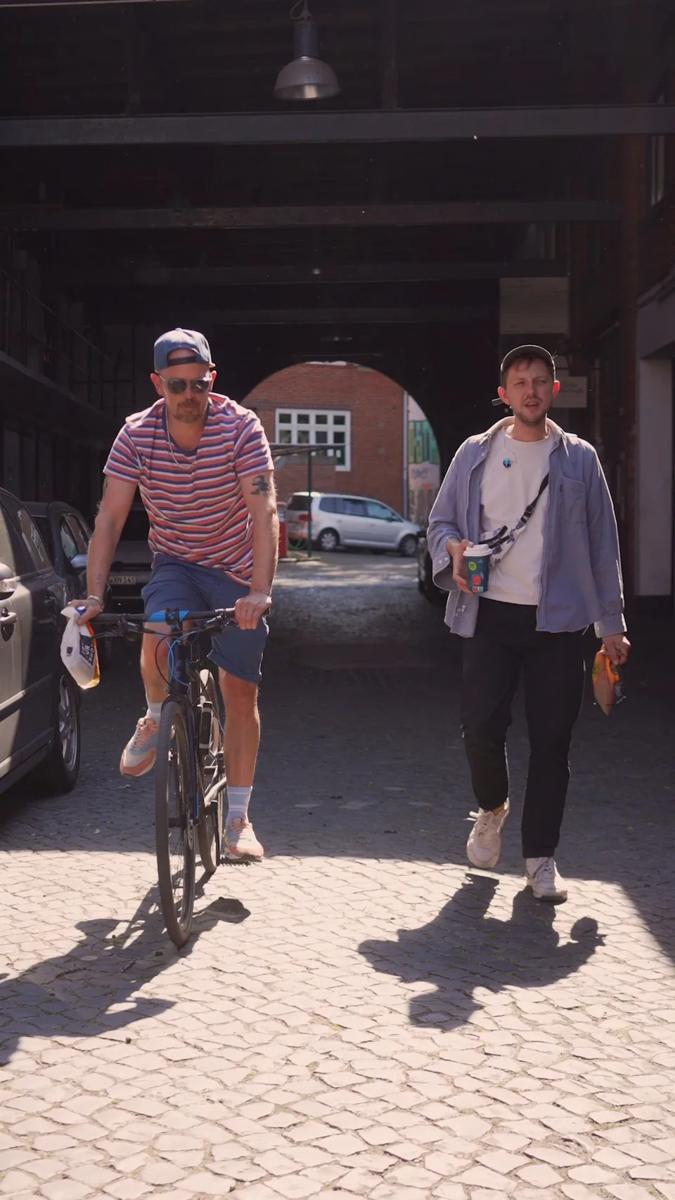 Zwei Männer gehen nebeneinander durch eine sonnige Hofdurchfahrt; einer hält ein Getränk.