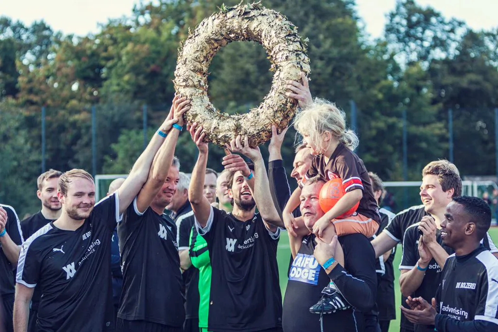Jubelnde Fußballer heben einen großen Kranz auf einem sonnigen Sportplatz; ein Kind sitzt auf den Schultern eines Mannes.