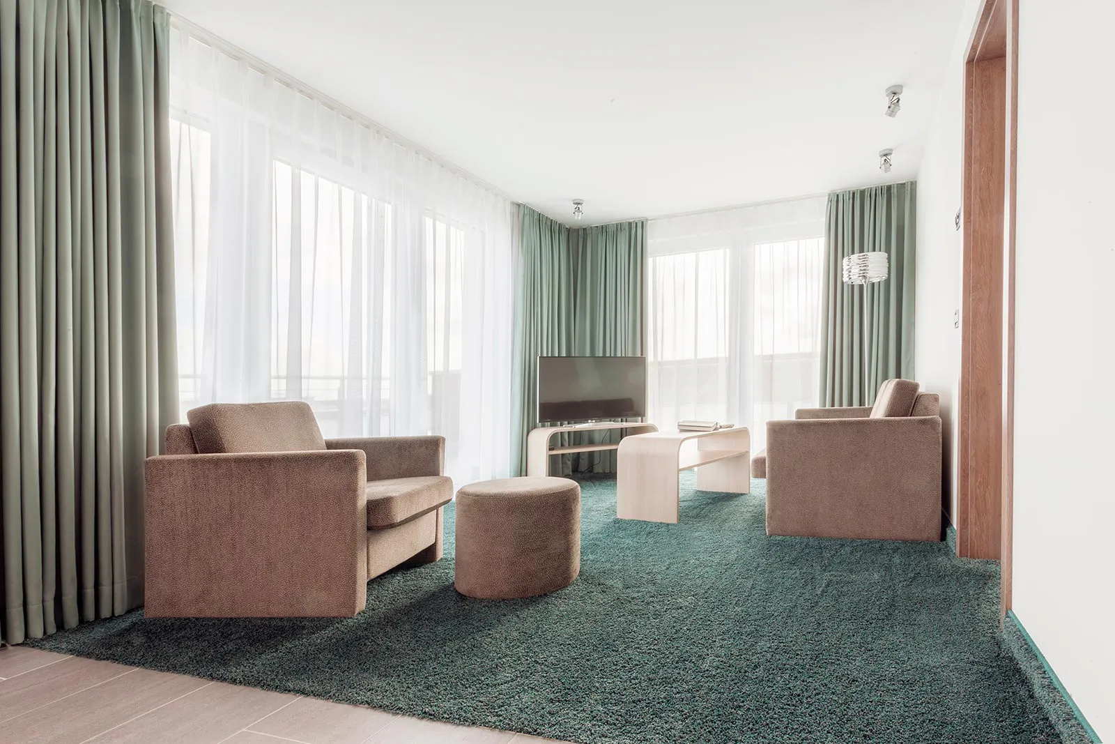 Heller Hotel-Wohnbereich mit beigen Sesseln, Hocker, Fernseher und grünem Teppich vor großen Fenstern.