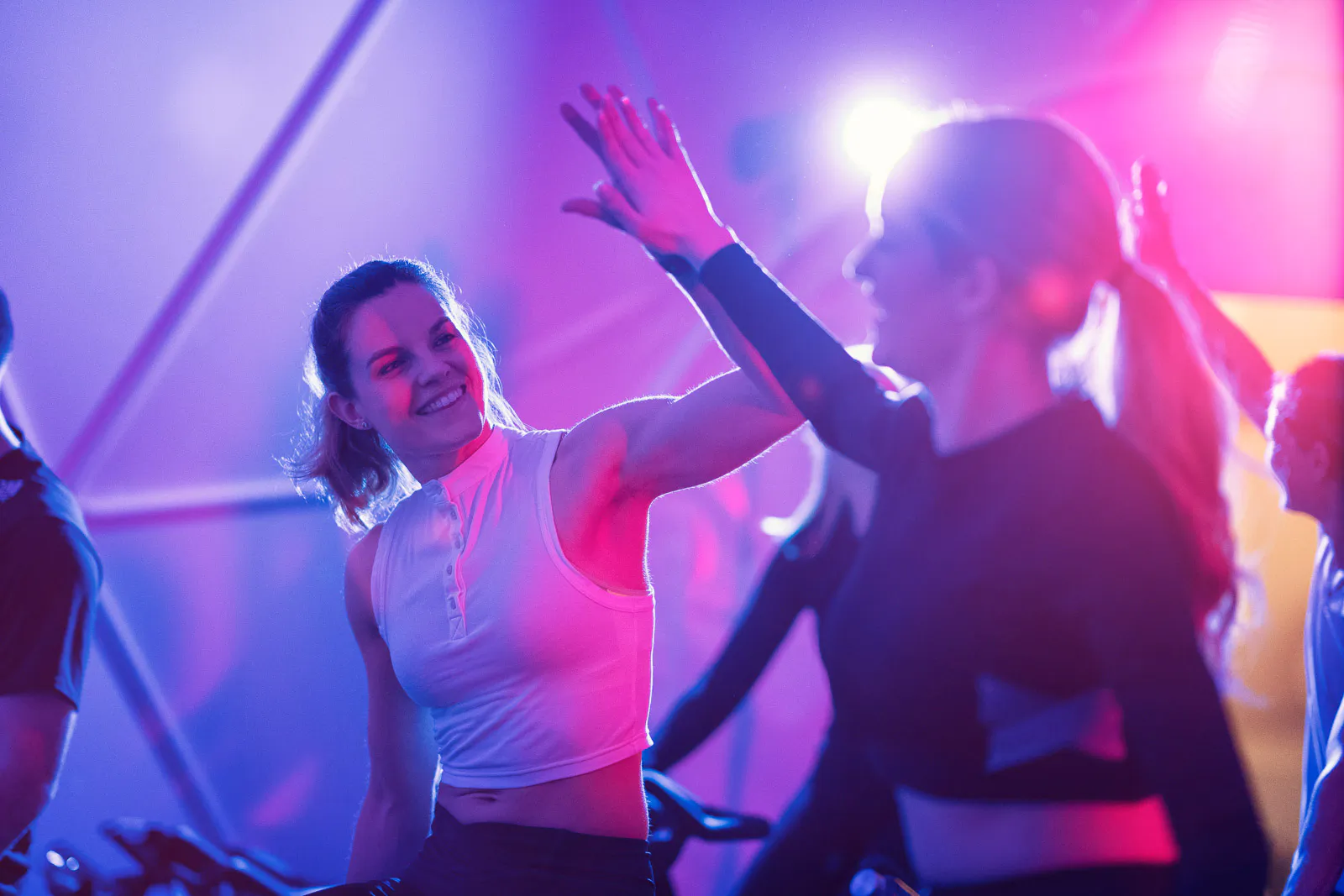 Zwei Frauen klatschen beim Gruppentraining im Fitnessstudio ab, beleuchtet von pinkem und blauem Licht.