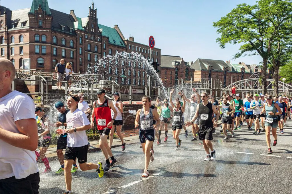 Läuferinnen und Läufer passieren bei Sonnenschein einen Wasserbogen auf einer Straße vor roten Backsteinhäusern.