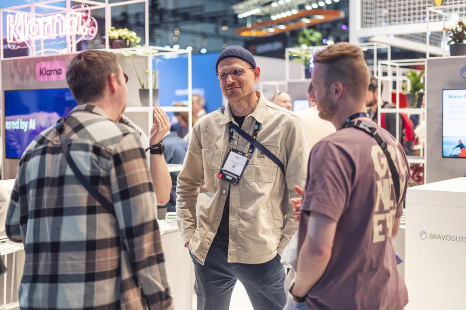 Drei Männer mit Messeausweisen unterhalten sich an einem hell beleuchteten Messestand mit Monitoren und Pflanzen.