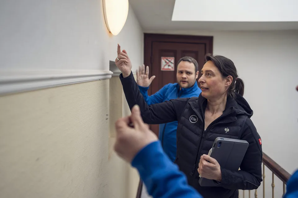 Frau mit Tablet zeigt im Treppenhaus auf eine leuchtende Wandleuchte, während ein Mann in Blau gestikuliert.
