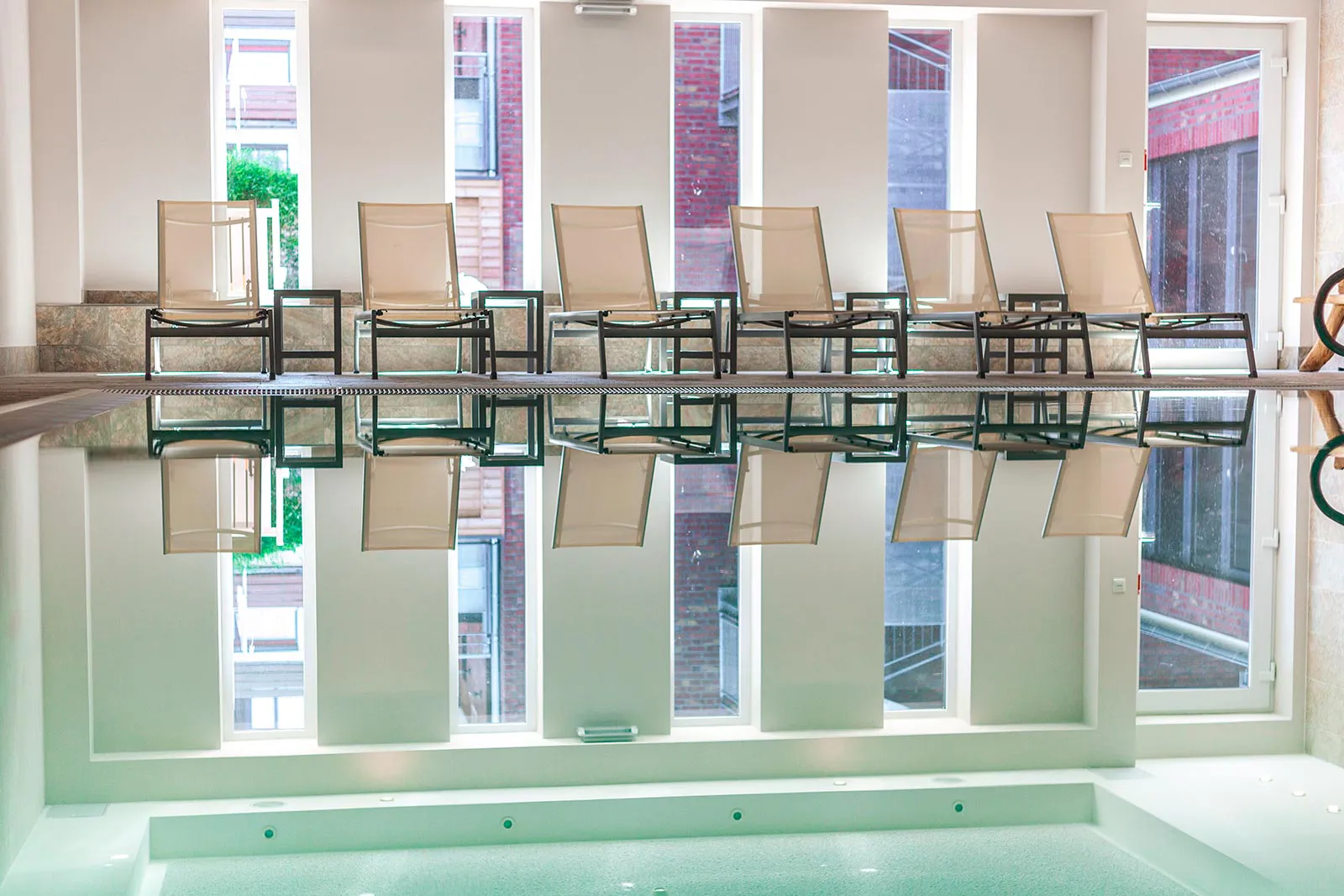 Hallenbad im Hotel mit sechs Liegestühlen vor hohen Fenstern, die sich im ruhigen Poolwasser spiegeln.