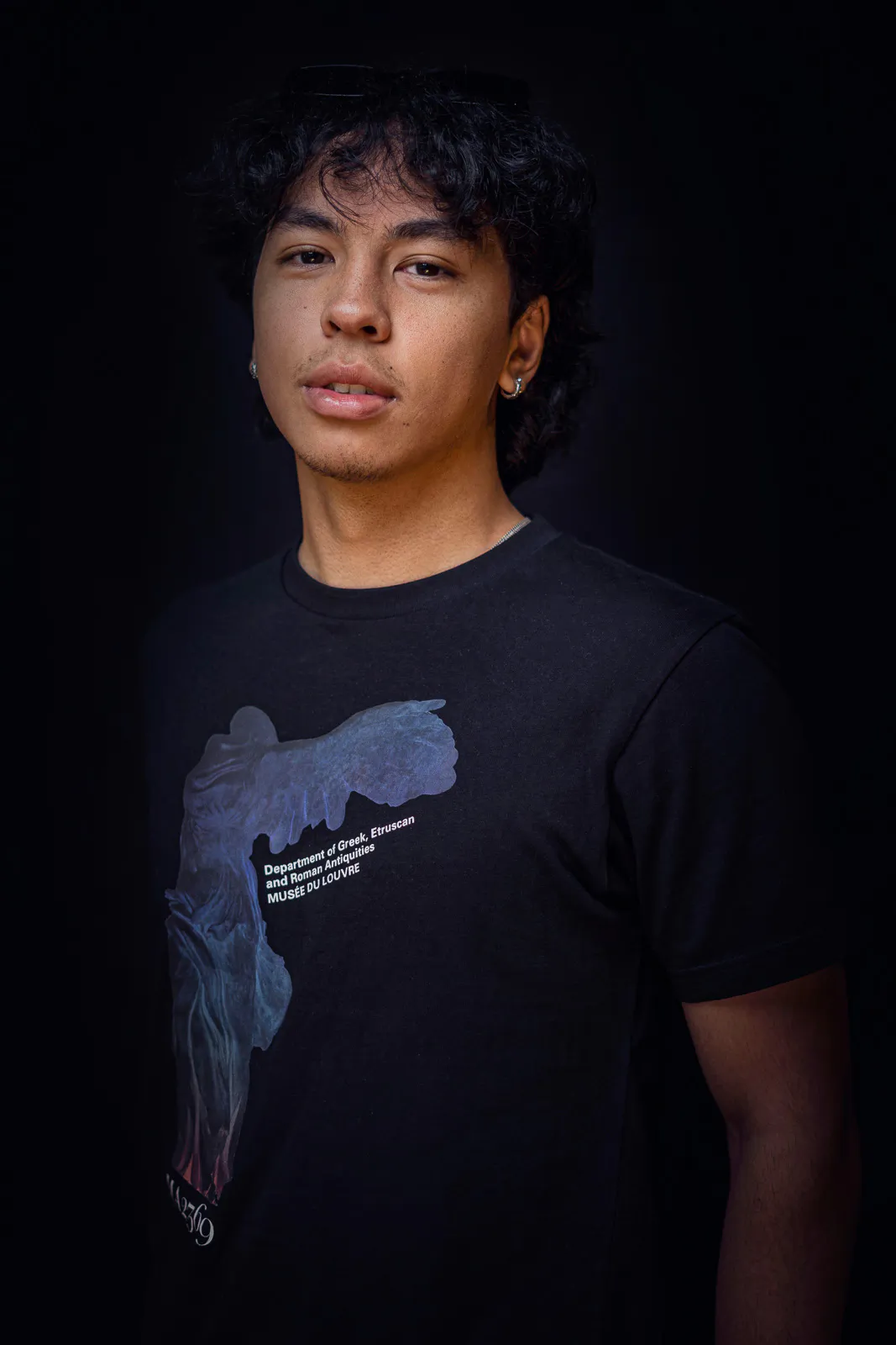Person mit dunklen Locken und bedrucktem schwarzem T-Shirt blickt vor schwarzem Hintergrund in die Kamera.