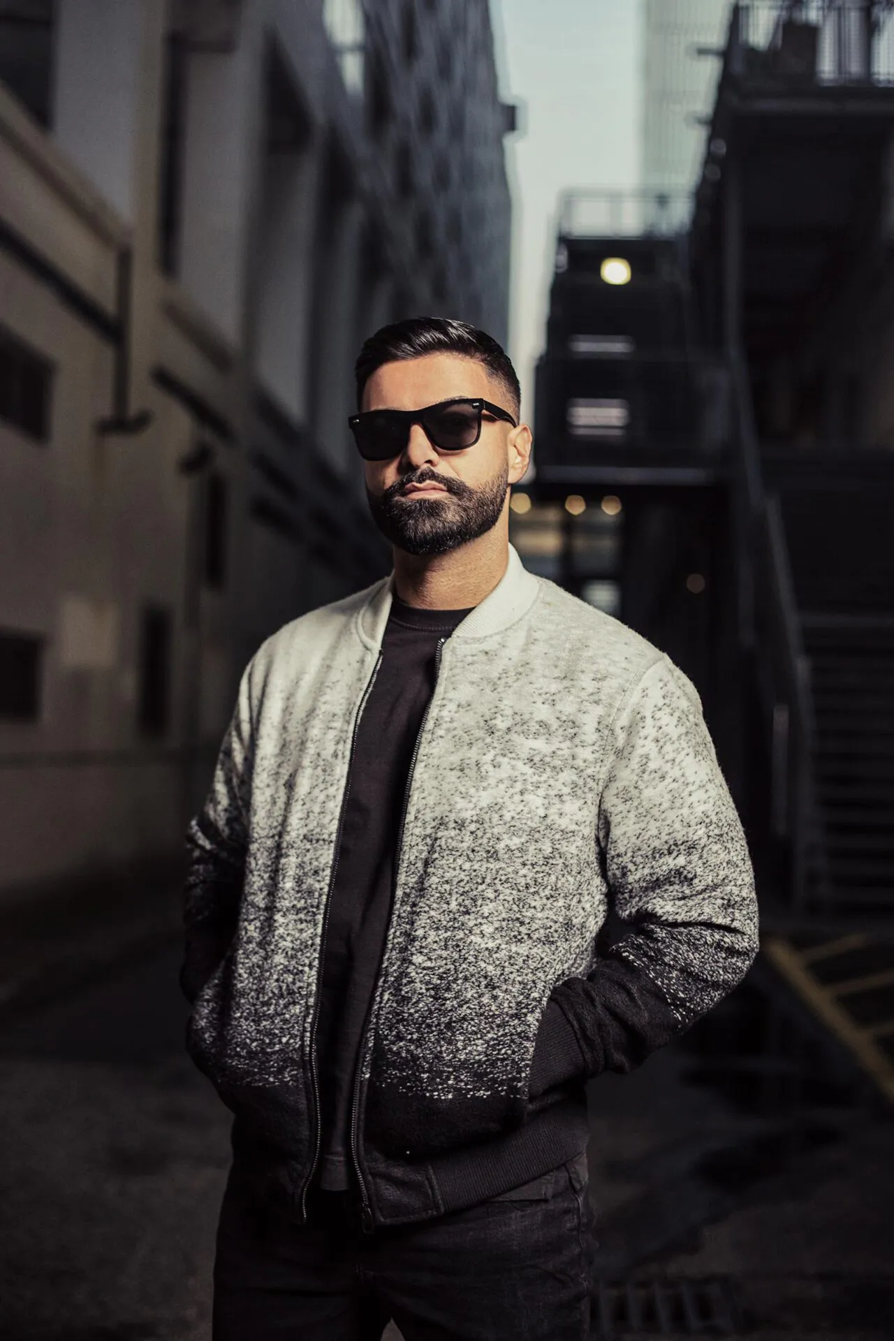 Mann mit Sonnenbrille und grauer Bomberjacke, Hände in den Taschen, im Tageslicht einer schmalen Gasse mit Metalltreppe.