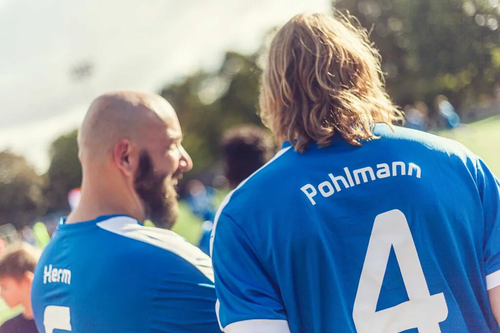 Zwei Männer in blauen Fußballtrikots stehen im Sonnenlicht auf einem Sportplatz, einer mit Rückennummer 4.