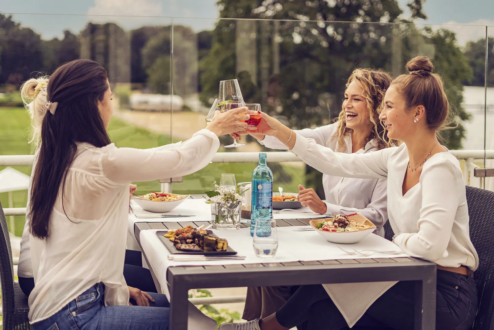 Frauen stoßen auf einer sonnigen Beachclub-Terrasse mit Getränken an, vor ihnen stehen Teller mit Essen.