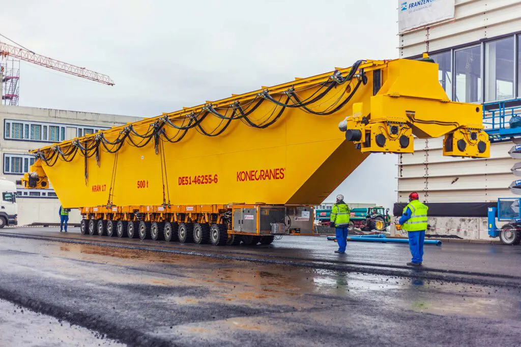 Gelber Konecranes-Kranträger auf Schwerlasttransporter, Arbeiter in Warnkleidung auf nassem Industriegelände.