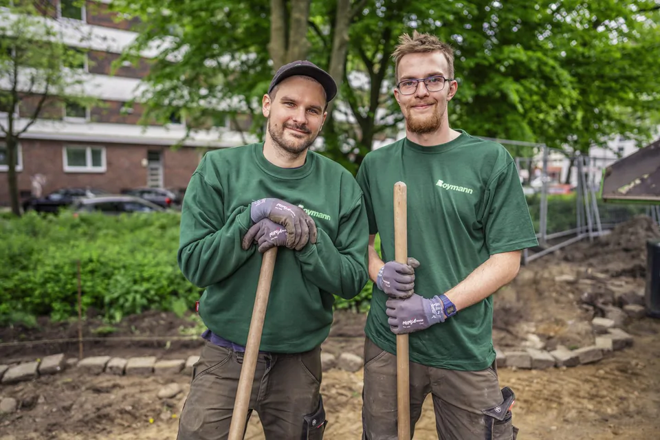 Zwei Männer in grüner Boymann-Arbeitskleidung halten Werkzeugstiele auf einer Gartenbaustelle im Tageslicht.