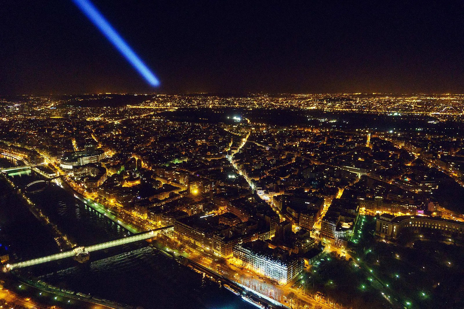 Nächtlicher Blick über Paris mit beleuchteten Straßen, Brücken über der Seine und blauem Lichtstrahl am Himmel.