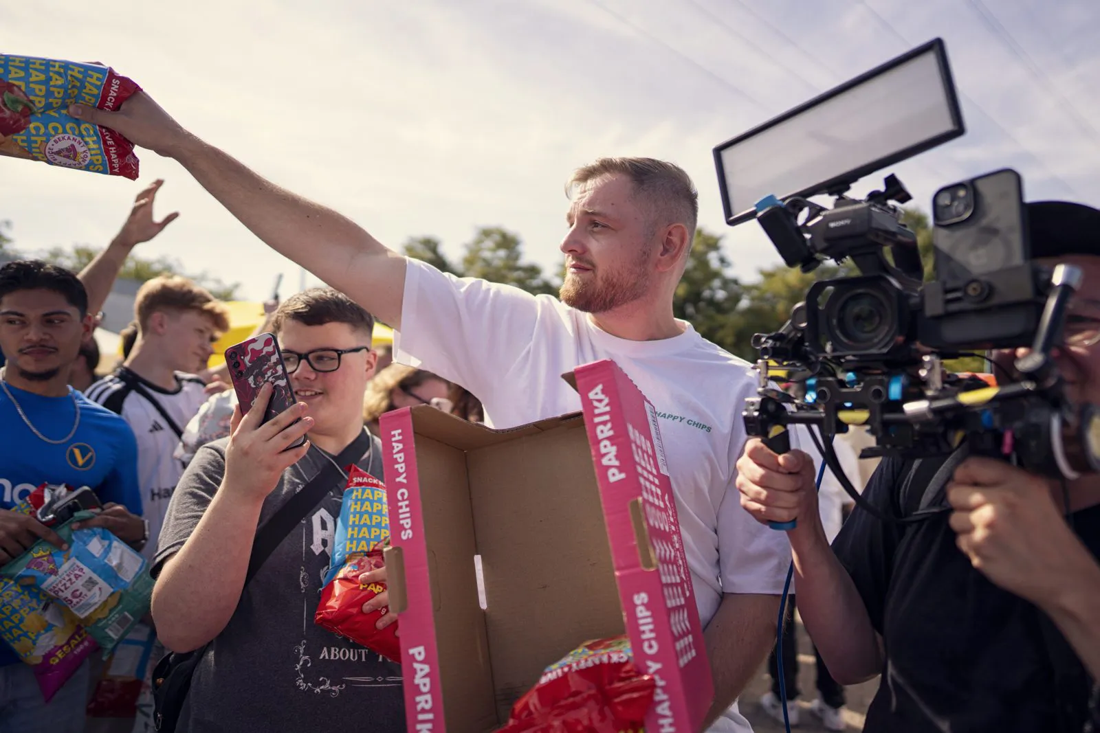 Mann verteilt Happy-Chips-Tüten an eine Menschenmenge im sonnigen Freien, daneben filmt ein Kameramann.
