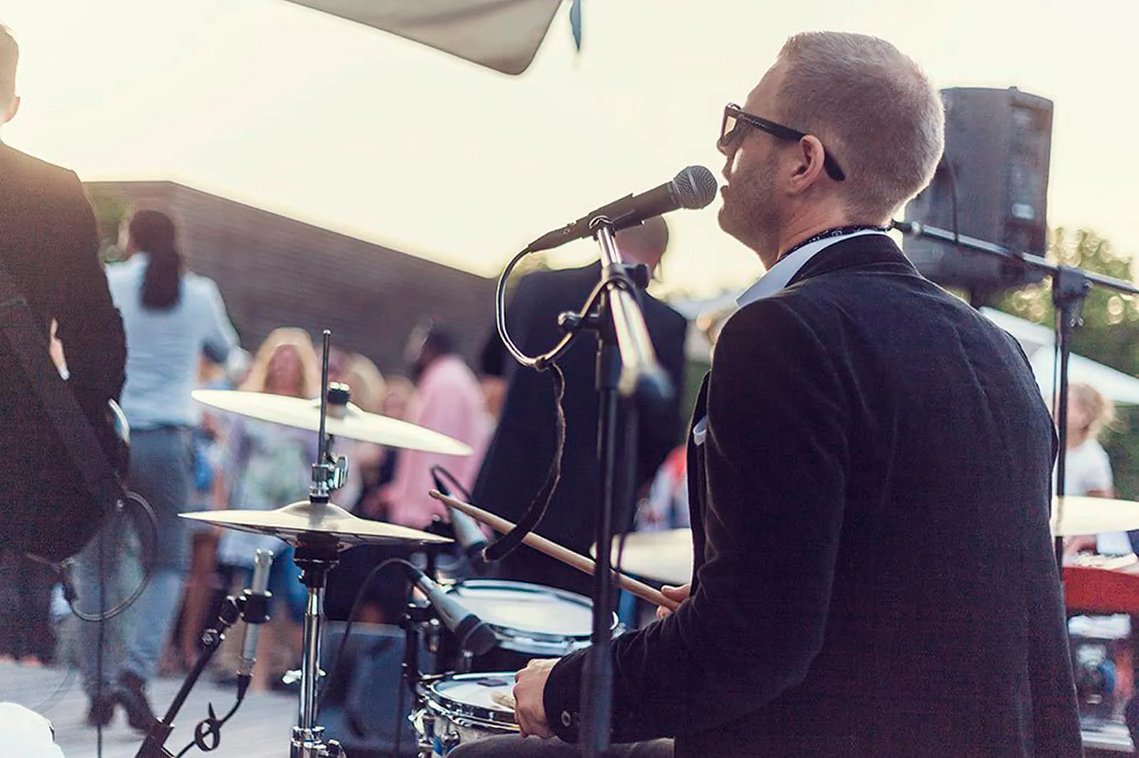 Schlagzeuger mit Sonnenbrille singt ins Mikrofon bei einem Open-Air-Event im warmen Gegenlicht, Gäste im Hintergrund.