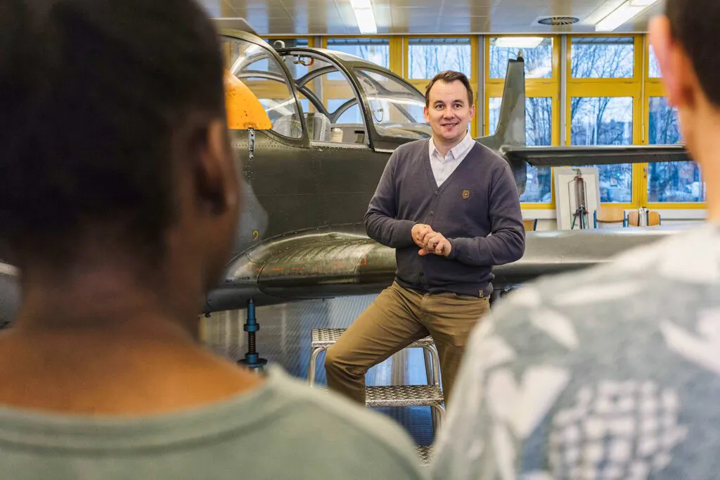 Ein Mann spricht vor zwei Personen neben einem Flugzeug in einer hellen Werkhalle mit großen Fenstern.