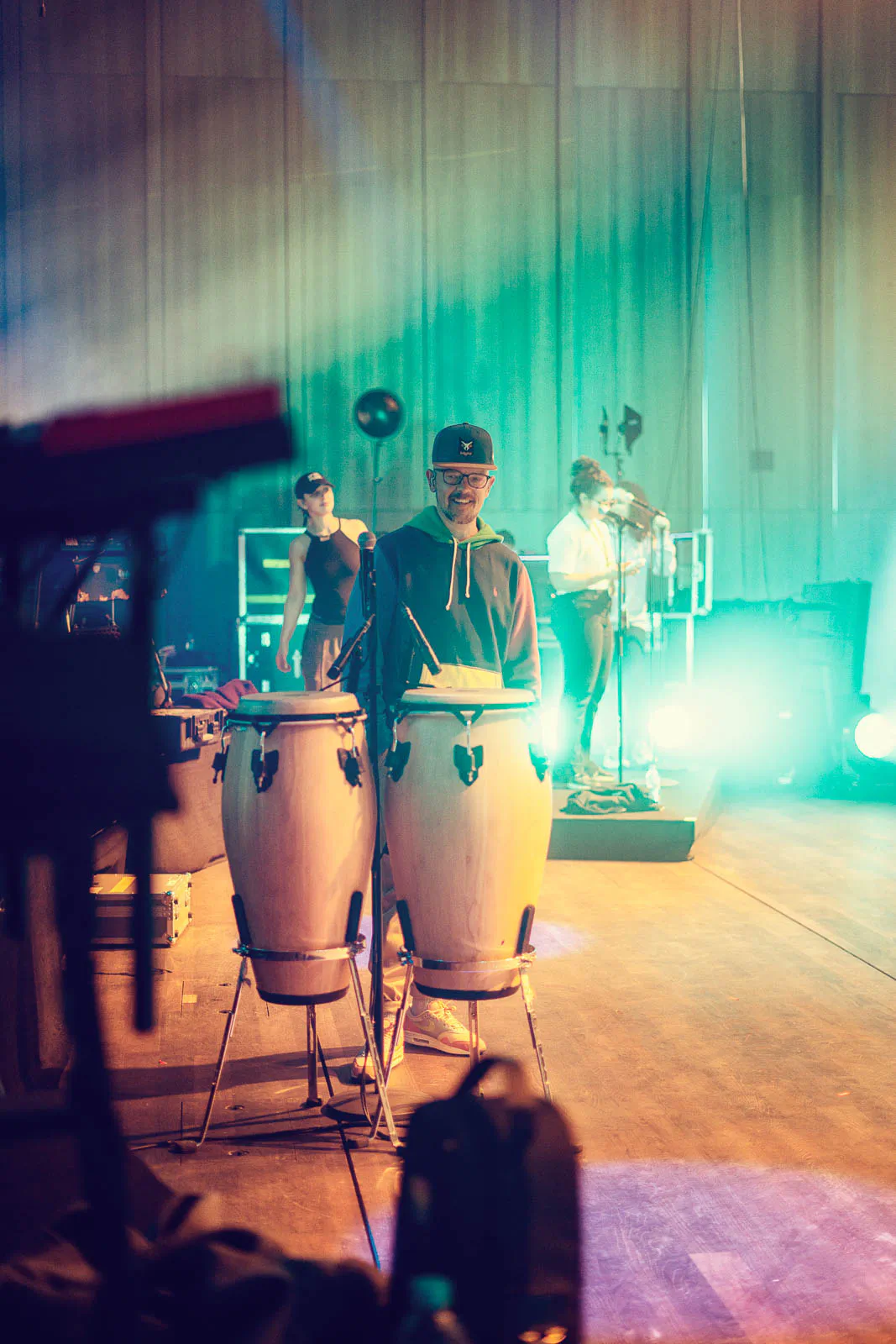 Perkussionist mit Kappe hinter zwei Congas auf einer Konzertbühne im grünen Licht, weitere Musiker im Hintergrund.
