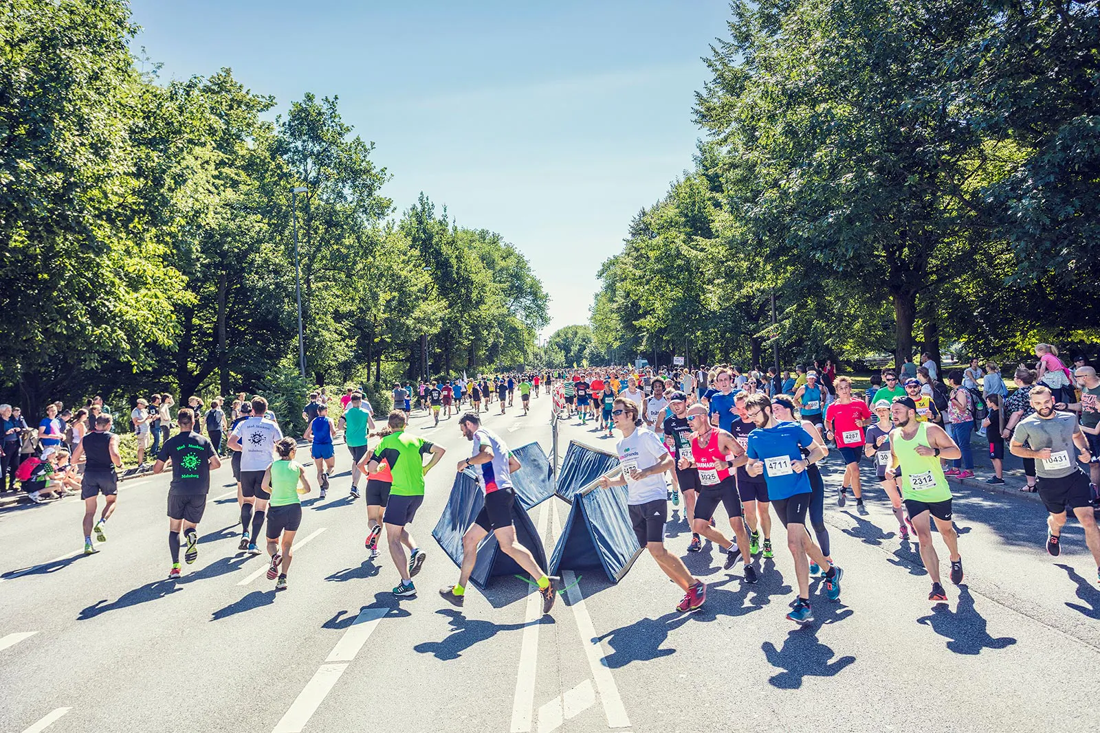 Läufer beim Hella Halbmarathon auf einer sonnigen, baumgesäumten Straße, einige tragen große schwarze Müllsäcke.