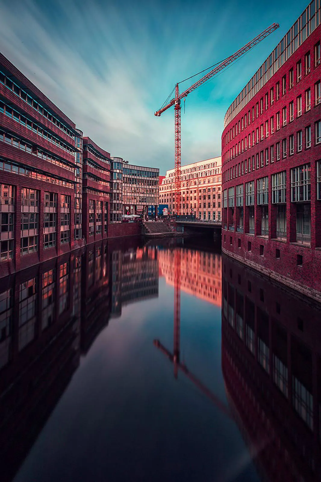 Rote Backsteingebäude und ein Baukran spiegeln sich im ruhigen Kanal unter einem blauen Himmel mit Wolkenschlieren.