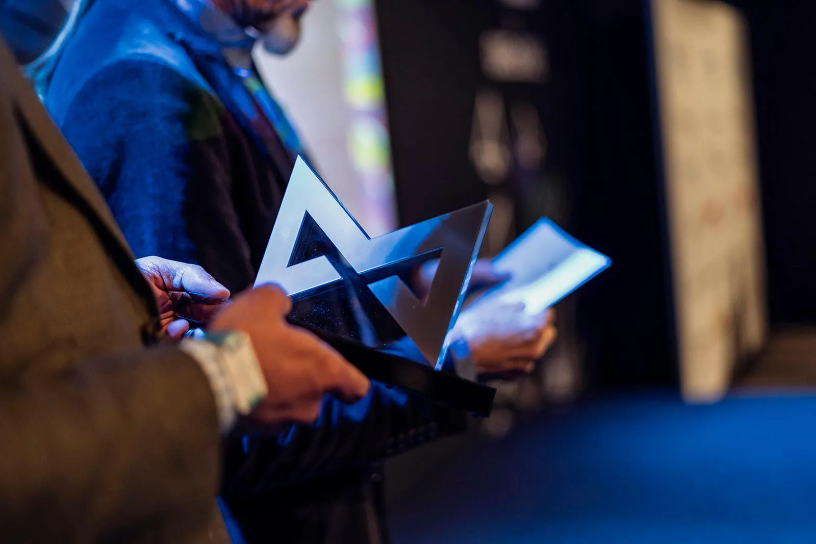 Personen halten eine geometrische MAX-Award-Trophäe und Unterlagen im blauen Veranstaltungslicht.