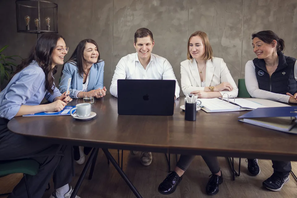 Fünf Personen sitzen bei einer Besprechung um einen Holztisch mit Laptop, Unterlagen und Kaffeetassen.