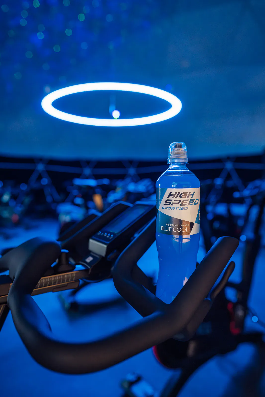 Blaue Sportgetränkflasche auf einem Spinningrad im Fitnessstudio unter blauem Licht und einer leuchtenden Ringleuchte.