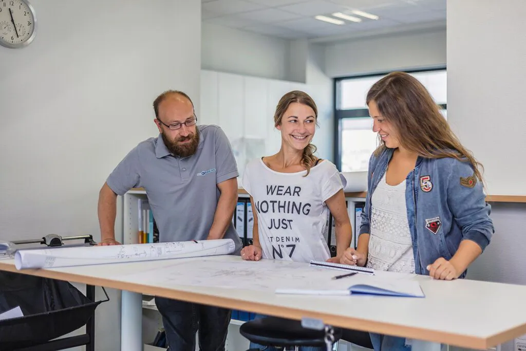 Ein Mann und zwei Frauen besprechen technische Zeichnungen an einem Tisch in einem hellen Büro.
