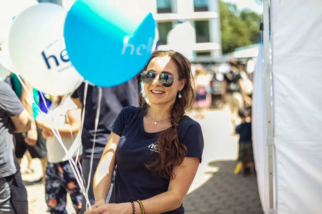 Lächelnde Frau mit Sonnenbrille hält weiße und türkise hella-Luftballons neben einem Zelt im Sonnenlicht.