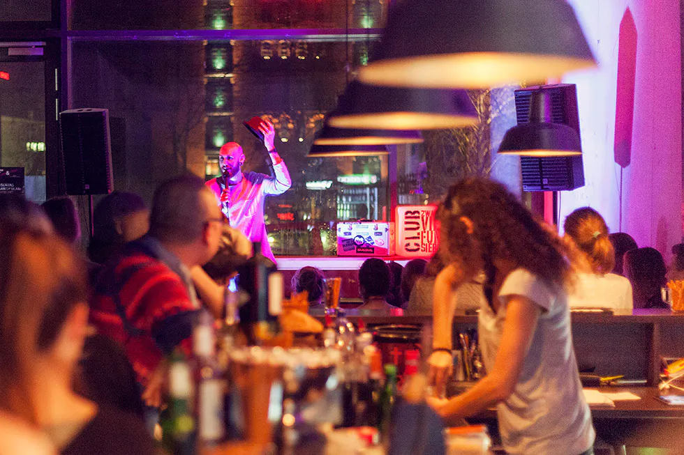 Dennis Grundt mit Mikrofon und erhobener Hand vor Publikum in einer Bar, beleuchtet von pinkem Bühnenlicht.