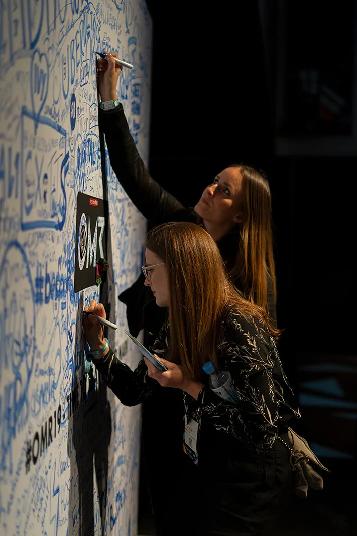 Zwei Frauen schreiben mit Stiften auf eine dicht beschriftete OMR-Wand in einem dunkel beleuchteten Innenraum.