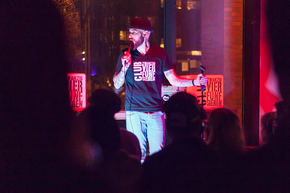 Dennis Grundt mit Hut und Mikrofon auf einer rot beleuchteten Stand-up-Bühne vor Publikum.