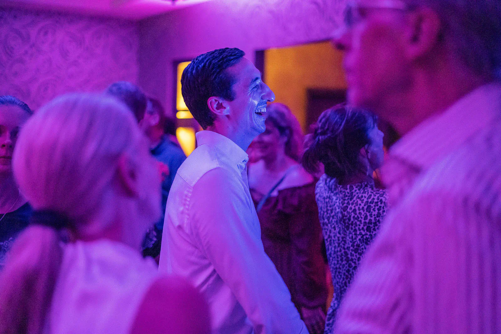 Lachender Mann im weißen Hemd zwischen Partygästen in einem Innenraum mit violettem Licht.
