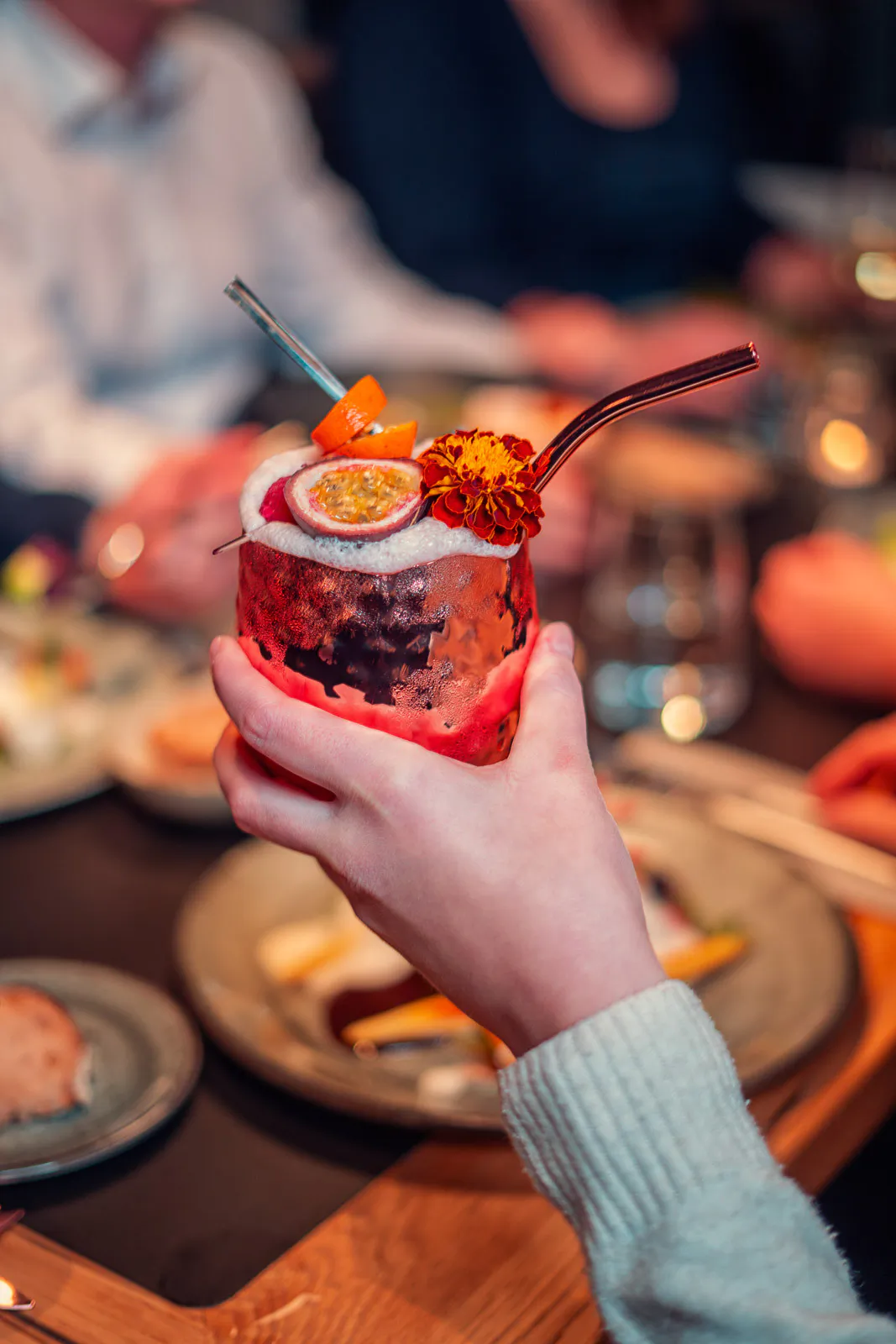 Hand hält Cocktail mit Passionsfrucht, Blüte und Metallhalmen am gedeckten Restauranttisch bei warmem Licht.