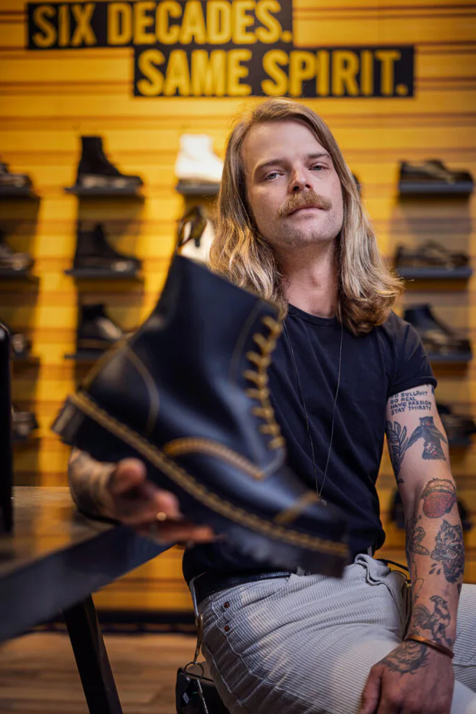 Langhaariger Mitarbeiter hält einen schwarzen Dr. Martens-Stiefel in die Kamera, vor gelber Schuhwand im warmen Licht.
