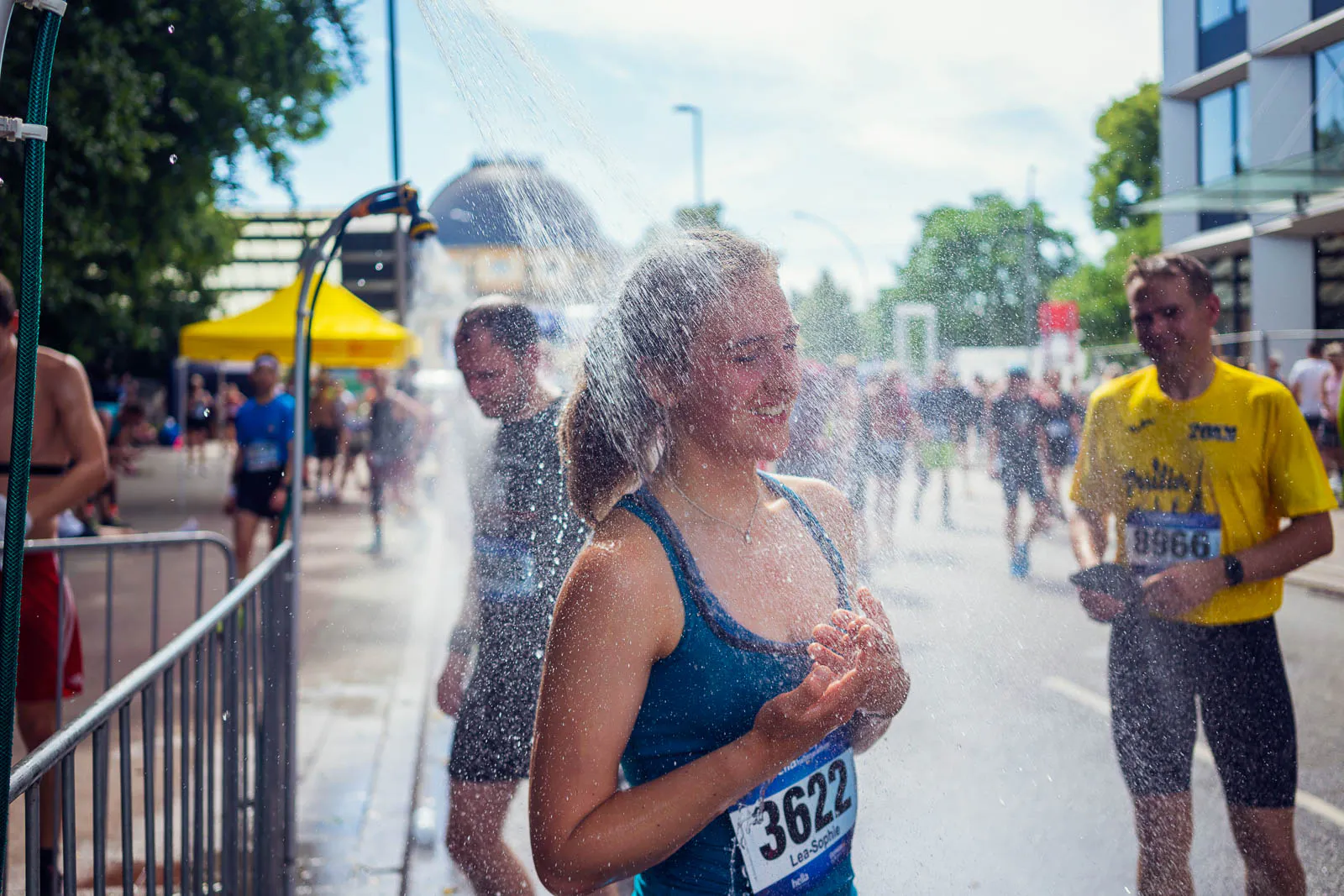 Läuferin im blauen Top kühlt sich beim Hella Halbmarathon unter einer Sprühdusche auf sonniger Straße ab.